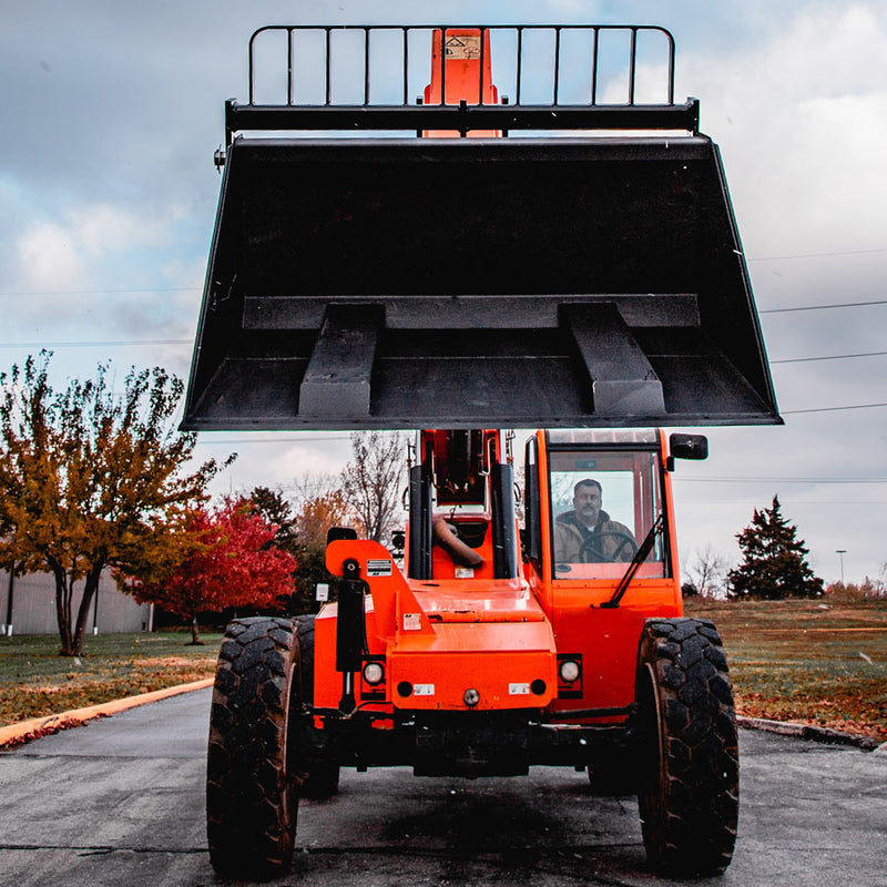 Arrow fork mounted bucket raised head-on above an orange telehandler, seen from below so both fork pockets and the bucket underside are in full view