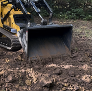 Arrow mini skid steer bucket mounted on a yellow Vermeer mini track loader, cutting into broken brown soil and rock with the bucket floor at grade