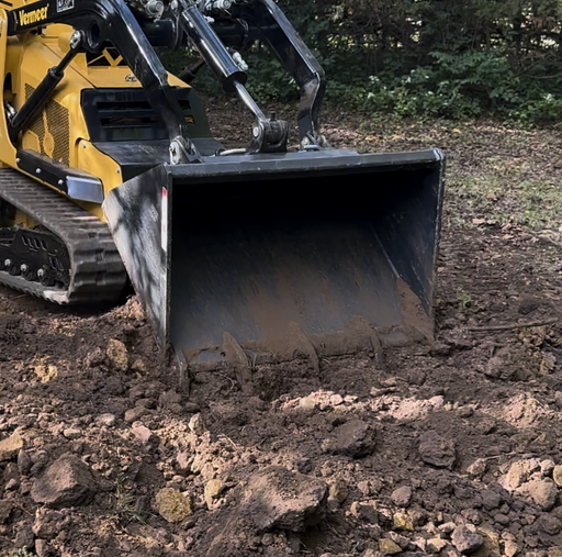 Arrow mini skid steer bucket mounted on a yellow Vermeer mini track loader, cutting into broken brown soil and rock with the bucket floor at grade