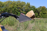 Arrow mini skid steer bucket carrying a large sandstone boulder on a loader arm in tall grass, the bucket tipped back with the plain cutting edge under the load