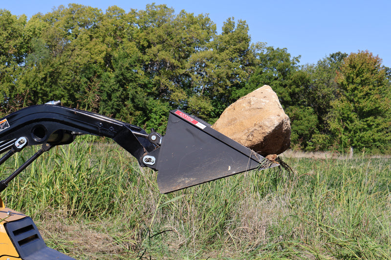 Arrow mini skid steer bucket carrying a large sandstone boulder on a loader arm in tall grass, the bucket tipped back with the plain cutting edge under the load