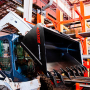 Arrow skid steer bucket mounted on a white skid steer inside a steel yard, shown fitted with optional bolt-on teeth along the cutting edge