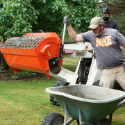 Worker pulling the manual discharge lever on the Hurricane mini mixer to empty wet mortar into a green wheelbarrow, with a second worker on the mini skid steer controls