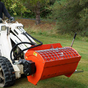Hurricane mini mixer carried on a white Ramrod Taskmaster mini skid steer on grass, showing the grate top, the mixing paddles beneath it and the black gear drive housing