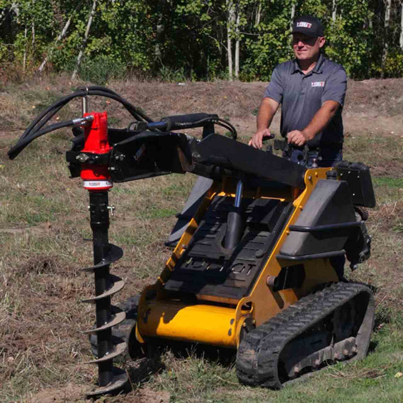 Operator standing at the controls of a yellow tracked mini skid steer carrying the Eterra auger drive and a black auger bit, in a grass field with birch woods behind