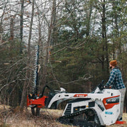 Operator on a white Bobcat MT100 mini track loader cutting into a standing pine with the sickle bar raised vertically against the trunk