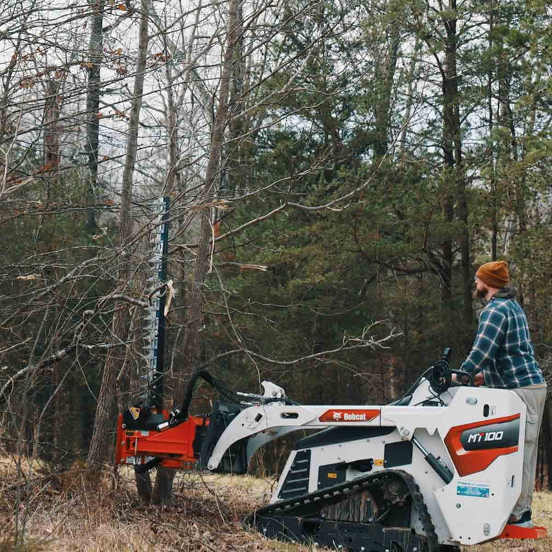 Operator on a white Bobcat MT100 mini track loader cutting into a standing pine with the sickle bar raised vertically against the trunk