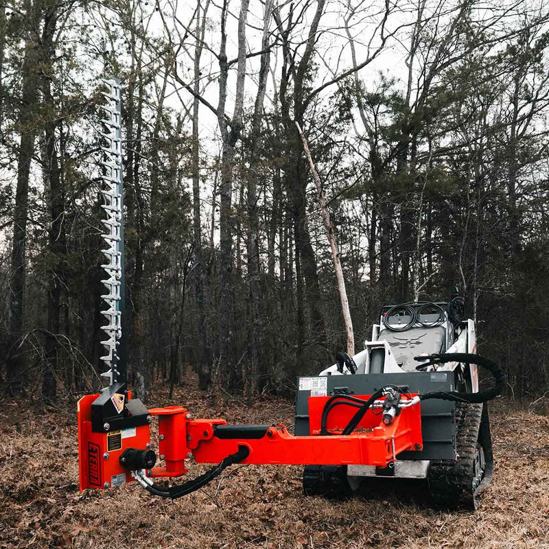 Side profile of the sickle bar mower on a white Bobcat mini track loader in bare woodland, showing the full orange arm, the pivot head and the vertical cutter bar