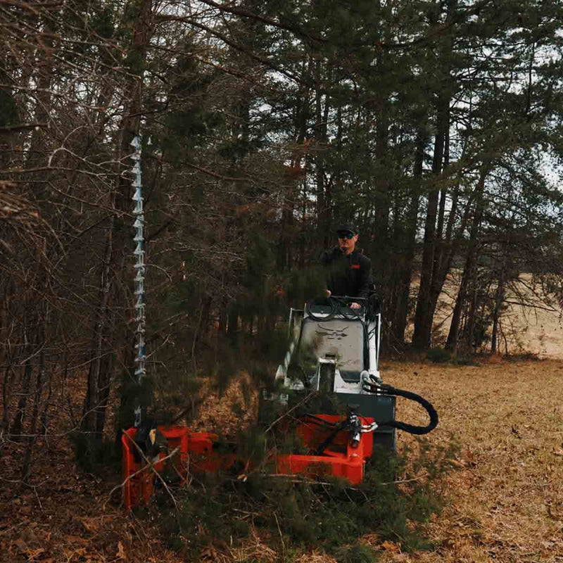 Sickle bar mower cutting into dense evergreen brush from a white Bobcat mini track loader, with the bar buried in the foliage