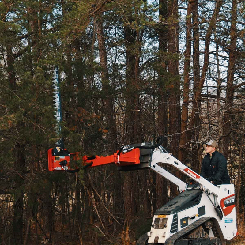 Sickle bar mower reaching up into pine branches with the arm extended from a white Bobcat MT100 mini track loader