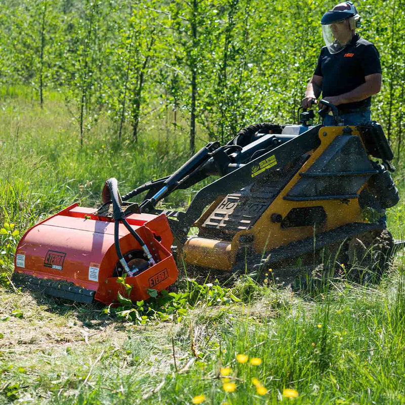 Operator in a face shield and ear defenders mowing rough grass and saplings with the Sidewinder flail mower on a yellow tracked mini skid steer