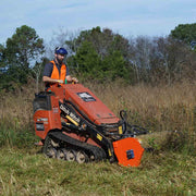Sidewinder flail mower mowing a rough field on an orange Ditch Witch SK750 mini skid steer, operated by a worker in a high-visibility vest and face shield