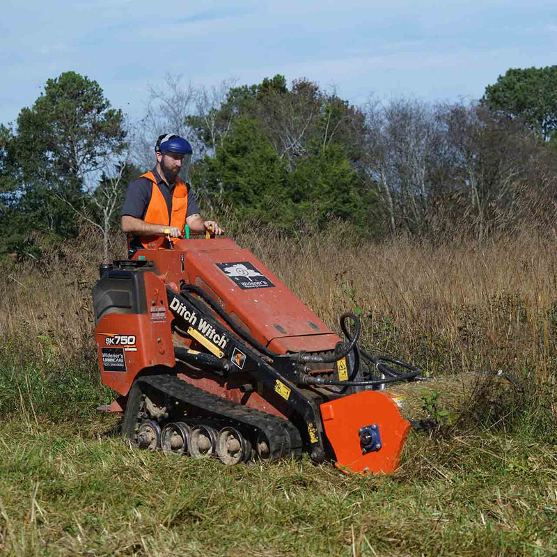 Sidewinder flail mower mowing a rough field on an orange Ditch Witch SK750 mini skid steer, operated by a worker in a high-visibility vest and face shield