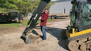 Operator standing beside a Glacier backhoe attachment mounted on a compact track loader in a gravel yard