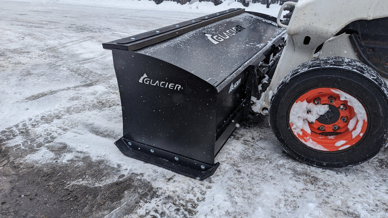 Glacier HD snow pusher mounted on a white skid steer clearing a snow-covered lot, side view