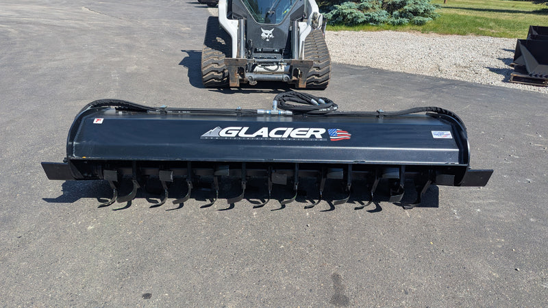 Glacier rotary tiller head-on behind a compact track loader, showing the full working width and the tine rotor