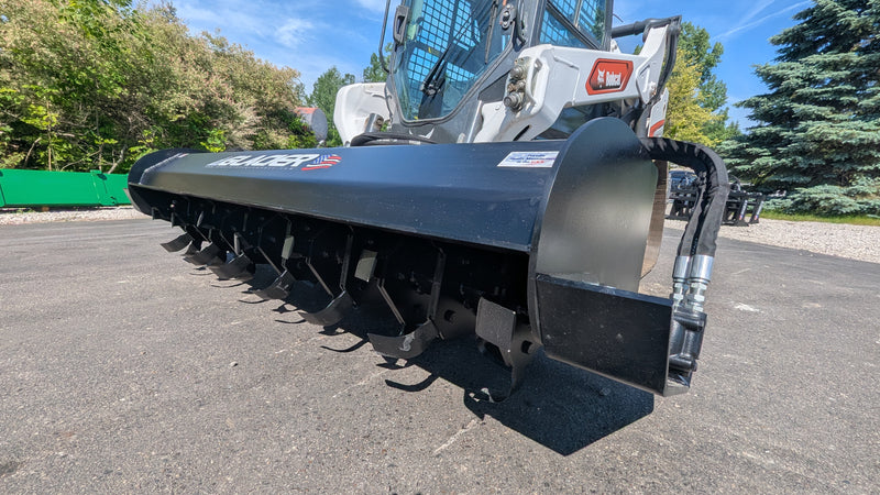 Glacier rotary tiller on a track loader, low angled view along the drum showing the tilling teeth and the side skid