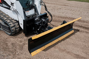 HitchDoc 6-Way Dozer Blade with a yellow top rail and black moldboard, mounted on a white Bobcat compact track loader on gravel, angled view showing the mount frame, hydraulic hoses and the bolted cutting edge