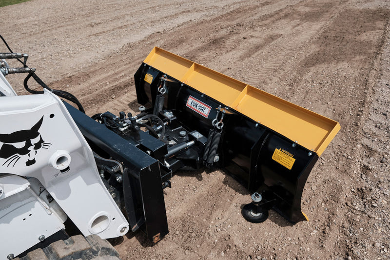 HitchDoc 6-Way Dozer Blade seen from above on gravel, showing the trip springs, the pivot cylinder, the yellow top rail and the caster at the blade end