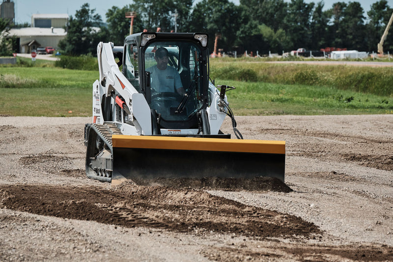 HitchDoc 6-Way Dozer Blade pushing gravel on a compact track loader, head-on view with the blade set straight and material rolling off both ends