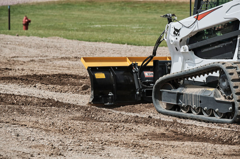 HitchDoc 6-Way Dozer Blade at work in gravel, close side view showing the moldboard, the trip spring and the blade angled to windrow material