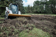 HitchDoc 6-Way Dozer Blade angled on a compact track loader, cutting and spreading dark soil over broken turf with the blade tilted to the cut