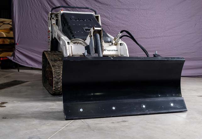 HitchDoc mini skid steer angle blade on a white Ingersoll Rand mini track loader indoors, head-on view of the one-piece rolled moldboard set straight, with the bolted reversible cutting edge along the bottom and the hydraulic hose running to the angling cylinder