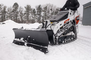 HitchDoc mini skid steer angle blade turned to the left on a white Bobcat MT100 mini track loader, three-quarter view of the one-piece rolled moldboard with the mount frame and hydraulic hoses behind it, in a snow-covered yard