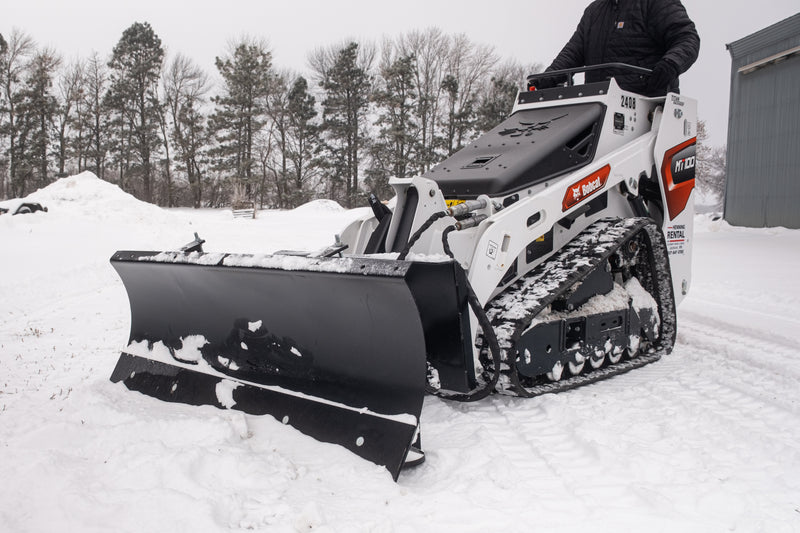 HitchDoc mini skid steer angle blade turned to the left on a white Bobcat MT100 mini track loader, three-quarter view of the one-piece rolled moldboard with the mount frame and hydraulic hoses behind it, in a snow-covered yard