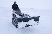 HitchDoc mini skid steer angle blade set straight and carrying snow along the moldboard, on a Bobcat MT100 walk-behind mini track loader with the operator at the controls on an open snow-covered lot
