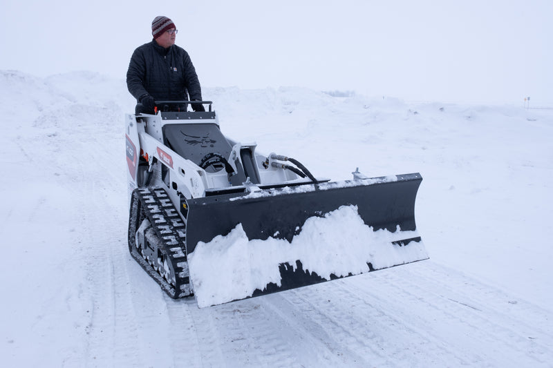 HitchDoc mini skid steer angle blade set straight and carrying snow along the moldboard, on a Bobcat MT100 walk-behind mini track loader with the operator at the controls on an open snow-covered lot