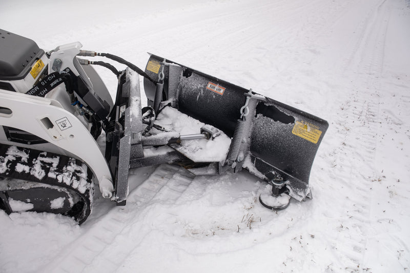 HitchDoc mini skid steer angle blade seen from behind on a Bobcat mini track loader, showing the A-frame between the two trip springs, the red and white KWIK-WAY decal and the warning decal on the moldboard back, and the skid shoe at the end