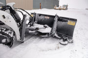 Rear three-quarter view of the HitchDoc mini skid steer angle blade showing the hydraulic angling cylinder across the push frame, both trip springs and the KWIK-WAY decal, with a shop building and trailers behind