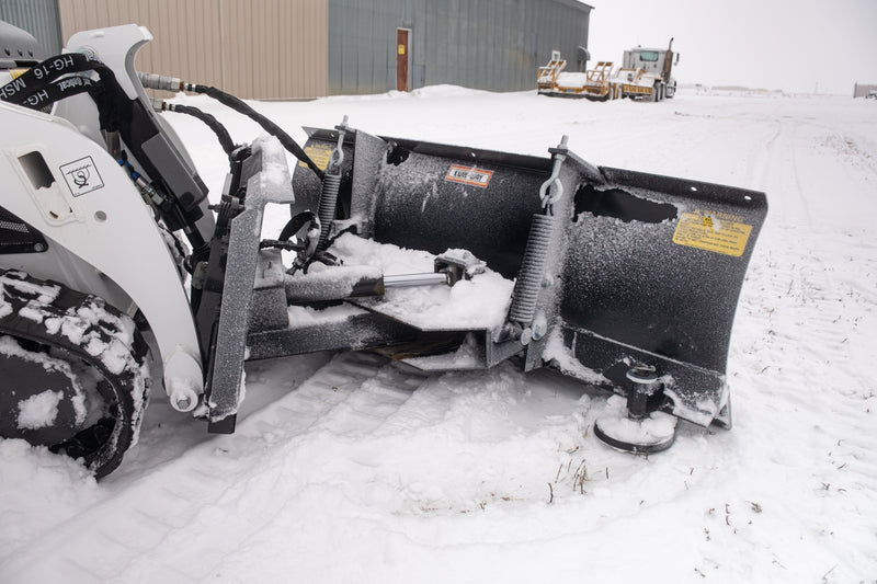 Rear three-quarter view of the HitchDoc mini skid steer angle blade showing the hydraulic angling cylinder across the push frame, both trip springs and the KWIK-WAY decal, with a shop building and trailers behind