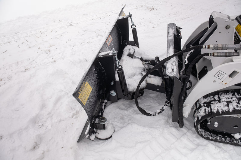 Close overhead view of the HitchDoc mini skid steer angle blade mount end, showing both trip springs, the angling cylinder, the quick connect hydraulic couplers, the yellow warning decal and the skid shoe below
