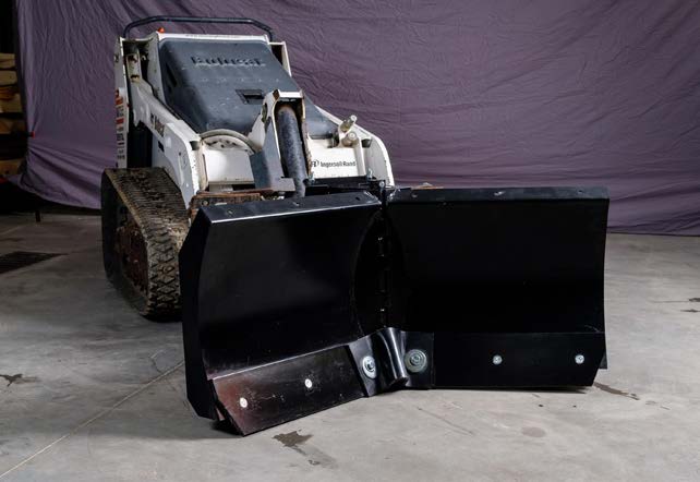 HitchDoc mini skid steer V-blade on a white Ingersoll Rand mini track loader indoors, both wings set forward in the scoop position on the centre hinge, with the bolted reversible cutting edges along the bottom of each wing