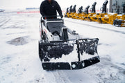 HitchDoc mini skid steer V-blade in the scoop position on a white Bobcat walk-behind mini track loader, both wings swept forward on the centre hinge with snow banked along the moldboards, in a snow-covered dealer yard with a row of snowblower attachments behind