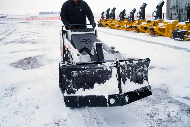 HitchDoc mini skid steer V-blade in the scoop position on a white Bobcat walk-behind mini track loader, both wings swept forward on the centre hinge with snow banked along the moldboards, in a snow-covered dealer yard with a row of snowblower attachments behind