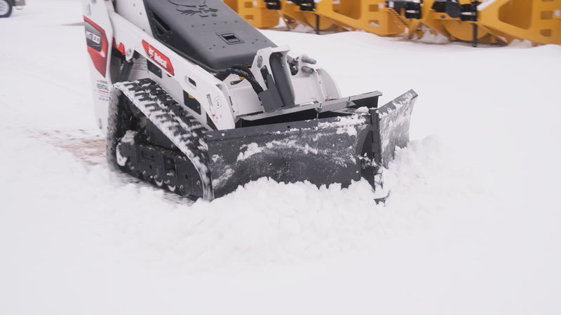 HitchDoc mini skid steer V-blade working on a Bobcat MT100 mini track loader, wings set offset on the centre hinge to cast snow to one side, with the bolted cutting edge riding down to bare ground