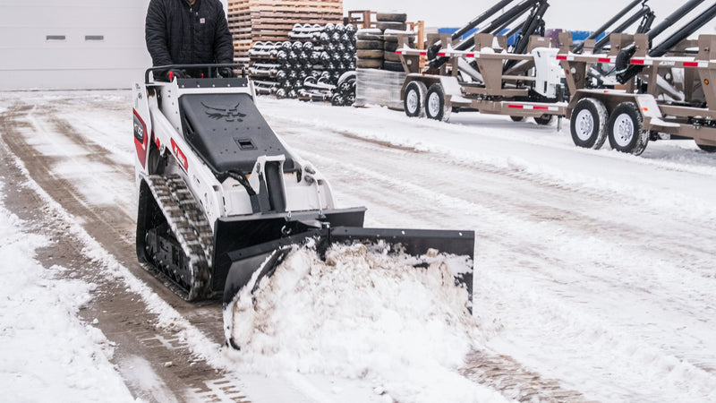 HitchDoc mini skid steer V-blade angled to windrow snow to the right on a Bobcat MT100 mini track loader, the operator walking behind at the controls and a cleared lane opening down to the surface