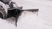 Close view of the HitchDoc mini skid steer V-blade on a mini track loader, showing the centre hinge, the trip spring, the mount frame and snow rolling off the leading wing