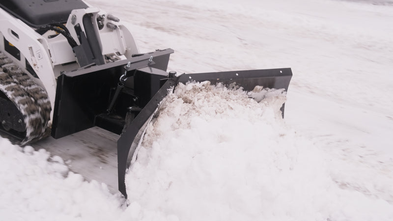 Close view of the HitchDoc mini skid steer V-blade on a mini track loader, showing the centre hinge, the trip spring, the mount frame and snow rolling off the leading wing