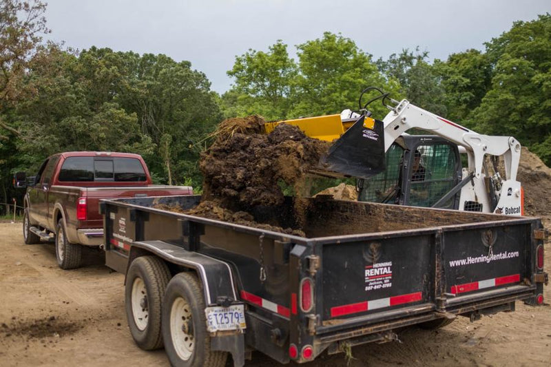 HitchDoc Push-Off Bucket carrying a heaped load of clay and sod over the side of a dump trailer, the yellow extractor just starting its stroke