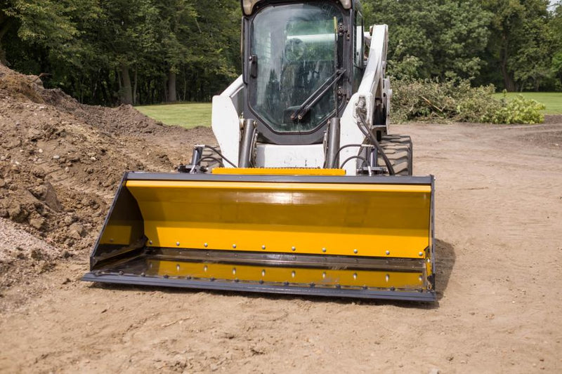 HitchDoc Push-Off Bucket seen head-on from the front on a white Bobcat skid steer, the yellow extractor blade across the back of the empty bucket and the bolted cutting edge resting on packed dirt