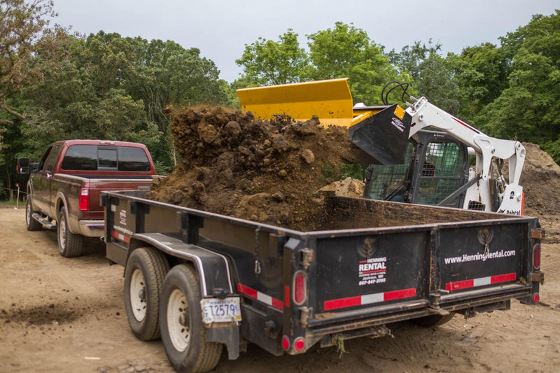 HitchDoc Push-Off Bucket ejecting a full load of heavy wet dirt into a black dump trailer, the yellow extractor blade extended and the load leaving the bucket without the machine tipping