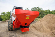 HitchDoc Sandpiper spreader mounted on a white Bobcat skid steer, rear three-quarter view of the red hopper carrying the Sandpiper script decal, with the discharge chute and spreader disc below and a sand pile alongside