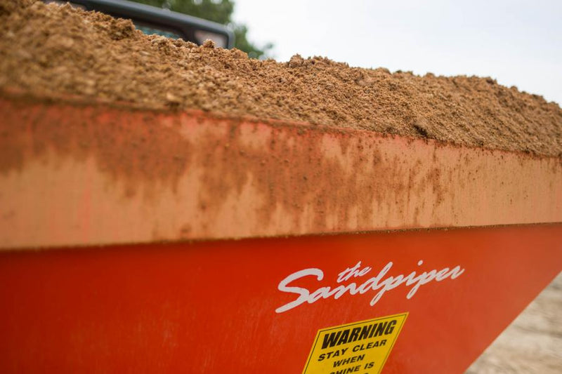 Close view of the HitchDoc Sandpiper hopper loaded level with damp sand, the Sandpiper script decal and the yellow warning decal on the red side wall