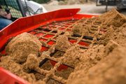 Lumps of damp sand resting on the HitchDoc Sandpiper protective grate as it screens material into the red hopper, with the skid steer and operator behind