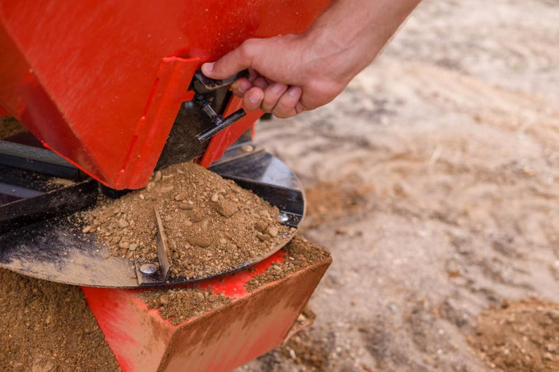A hand setting the manual gate lever at the base of the HitchDoc Sandpiper hopper, with the steel spreader disc below carrying a cone of sand