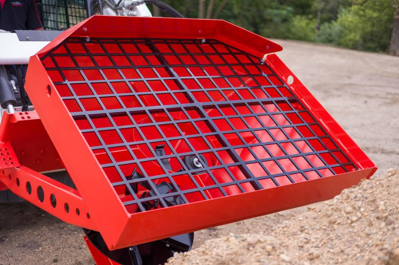 Top view of the HitchDoc Sandpiper protective grate bolted across the red hopper, its square openings showing the spreader disc and agitator below, mounted on a white skid steer